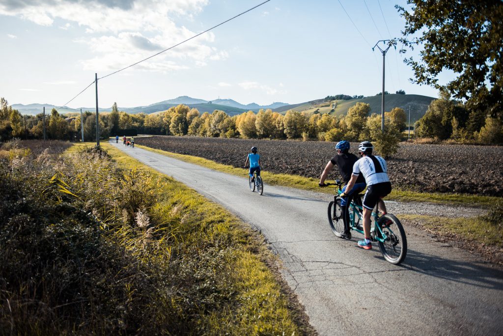Dos ciclistas en tándem detrás de otro ciclista recorren un camino asfaltado rodeado de árboles otoñales.