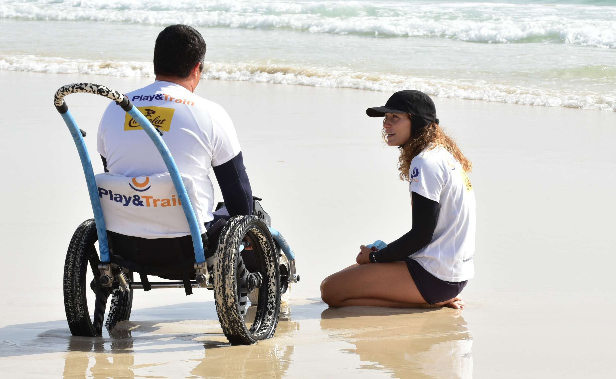 Un hombre sentado en una silla de ruedas para la playa junto a su monitora de surf
