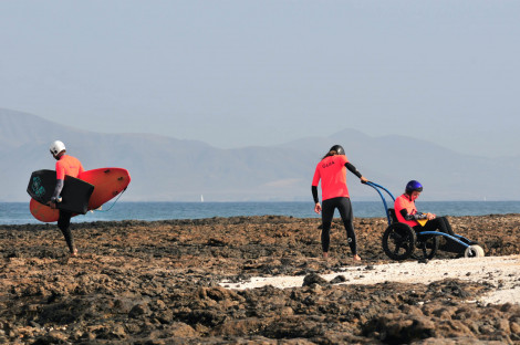 Tres personas avanzando por una playa rocosa. Una de ellas sentada en una silla de ruedas para playa mientras otra la ayuda y la tercera con las tablas de surf.
